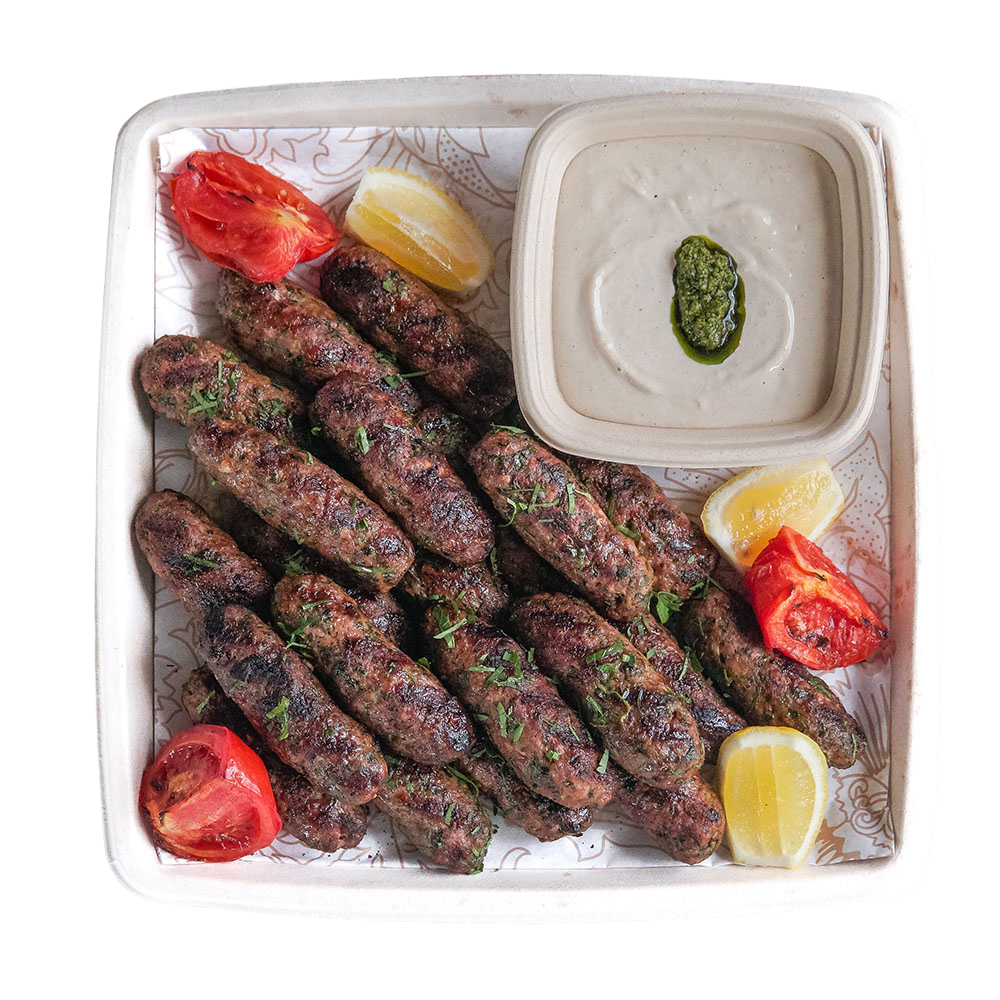 A white tray of grilled kebabs garnished with chopped herbs, with lemon wedges, roasted tomatoes, and a bowl of white sauce with a drizzle of green sauce.