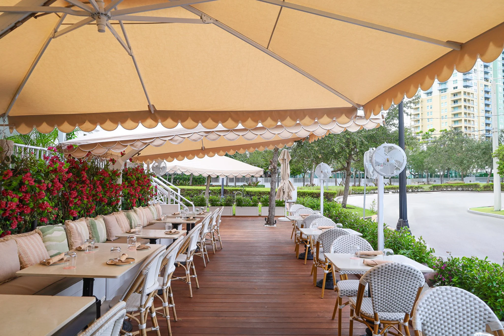 Outdoor restaurant patio with beige umbrellas, white chairs, and tables set for dining, surrounded by pink flowers and green trees.