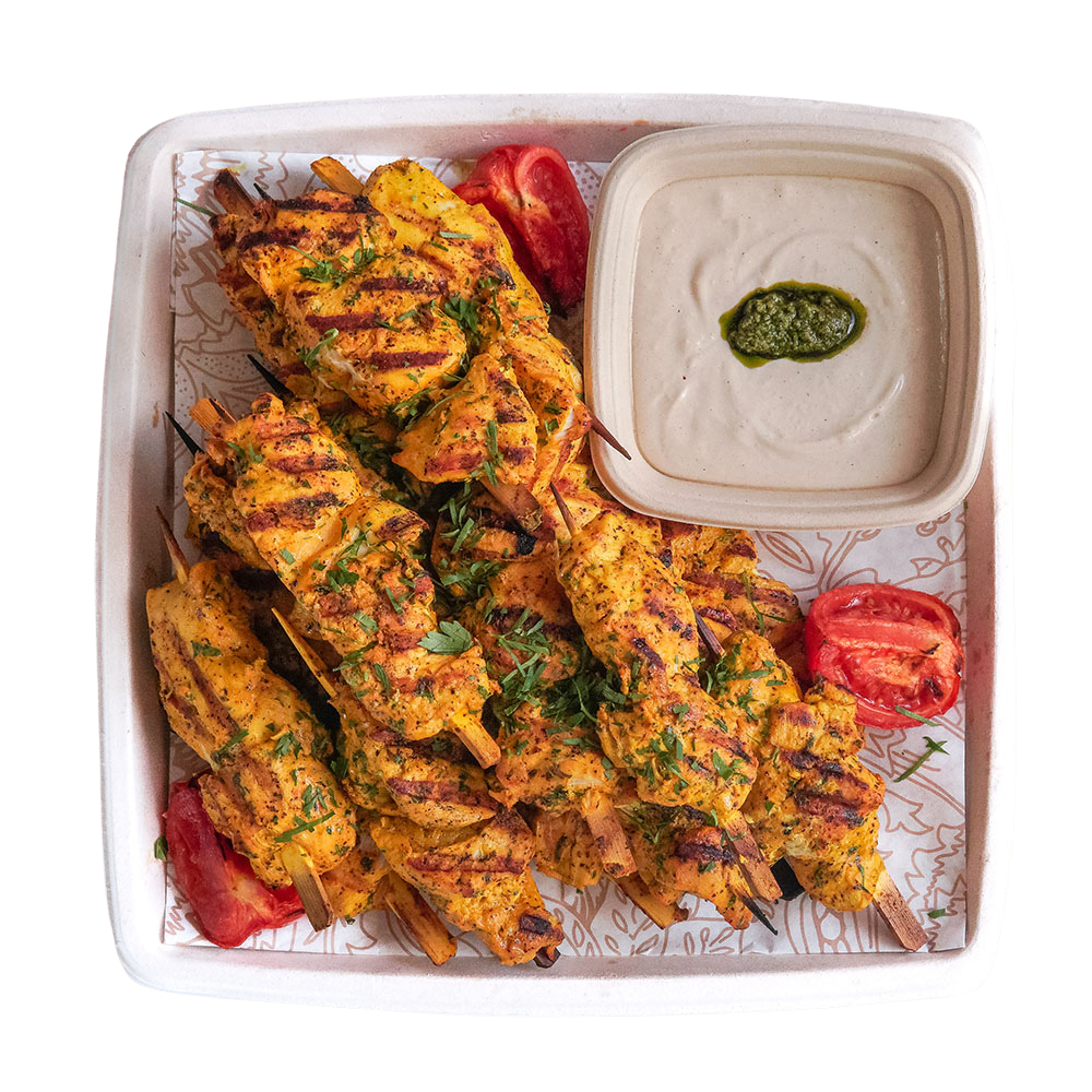 Dish with grilled chicken skewers garnished with herbs, sliced tomatoes, and a side of green sauce in a plastic container.