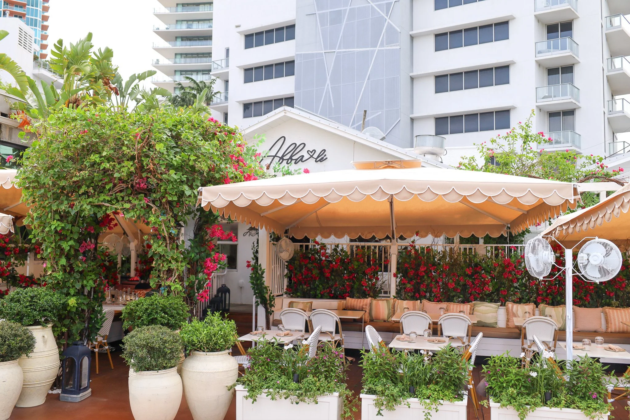 Outdoor patio at a restaurant with umbrellas, tables, chairs, and potted plants with a white building and high-rise apartments in the background.