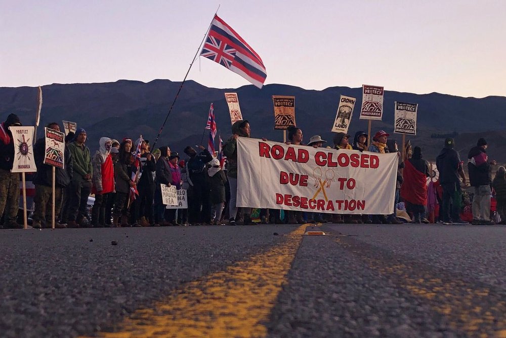  Demonstrators block a road on the Big Island’s  Maunakea to protest the construction of a giant telescope on land that  Native Hawaiians consider sacred, on August 5, 2019. (Image: Caleb Jones/AP via Vox https://www.vox.com/2020/3/30/21198011/hawaii