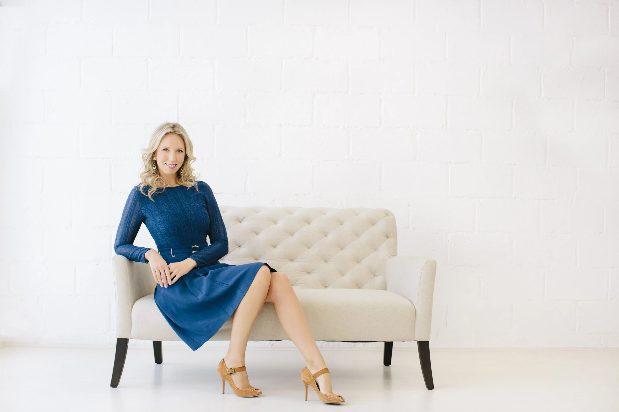 A woman in a blue dress sitting on a cream-colored tufted sofa in a bright, minimalistic room.