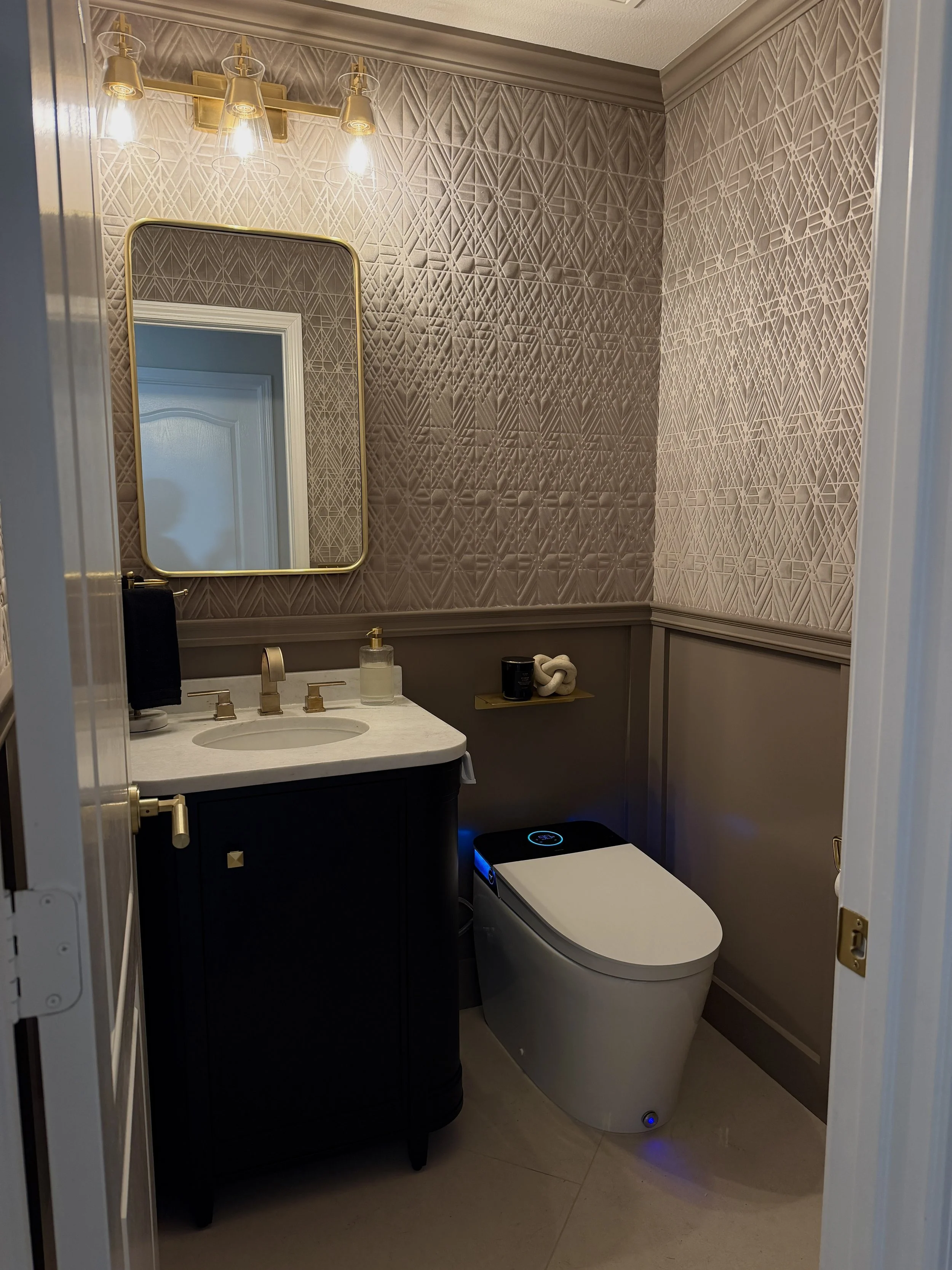 Transitional powder room with textured geometric wallpaper, a black vanity with a marble top, gold fixtures, and a smart toilet by Gin Treadwell Interiors in Santa Clarita.