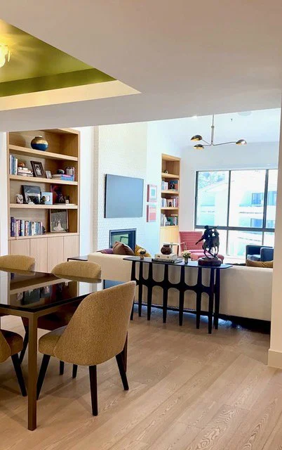 Living room with a wooden dining table, beige chairs, built-in shelves with books and decor, wall-mounted TV, white sofa, black side table, plants, large window with view outside.