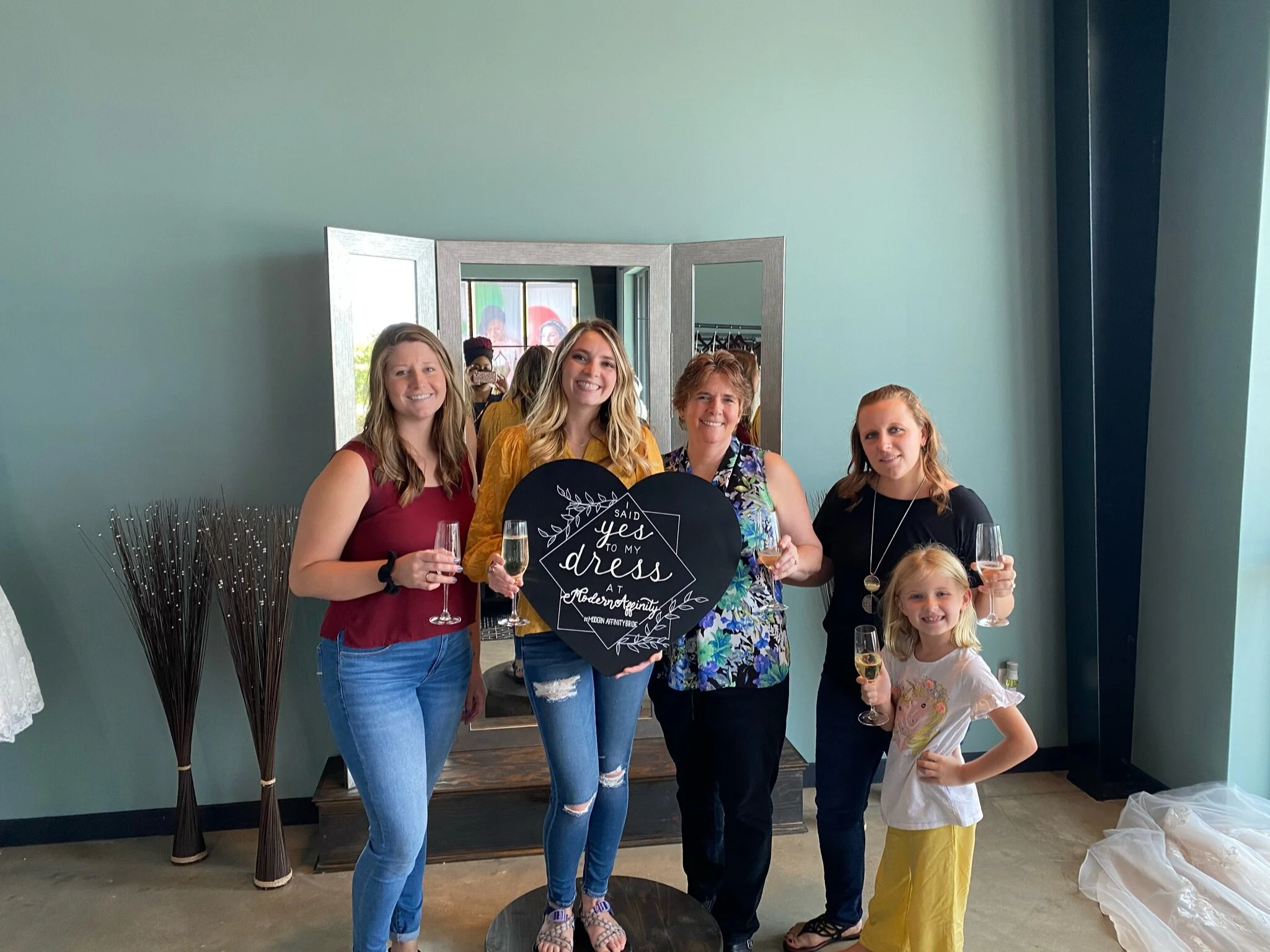 A group of five women and one young girl celebrating with champagne in a room with a teal wall. One woman holds a heart-shaped sign that says, 'Said Yes to My Dress at Modern Tuxedo.' They are smiling and posing for the photo.