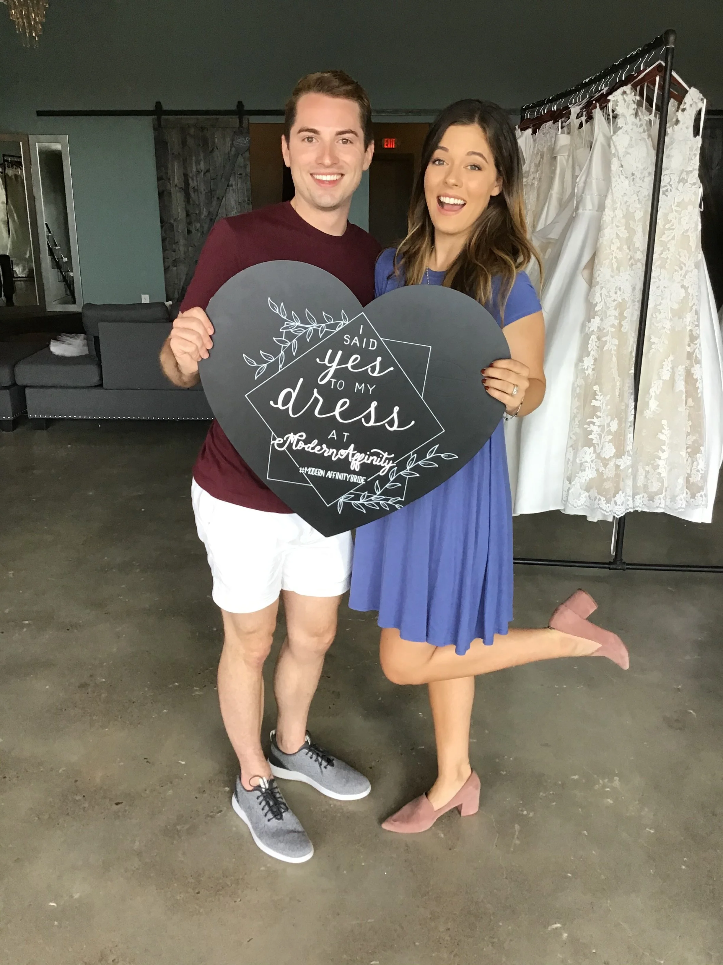 A young man and woman posing together indoors, holding a heart-shaped sign that reads 'I said yes to my dress a modern affinity'. The woman is wearing a blue dress and pink heels, while the man is wearing a maroon shirt, white shorts, and sneakers. T