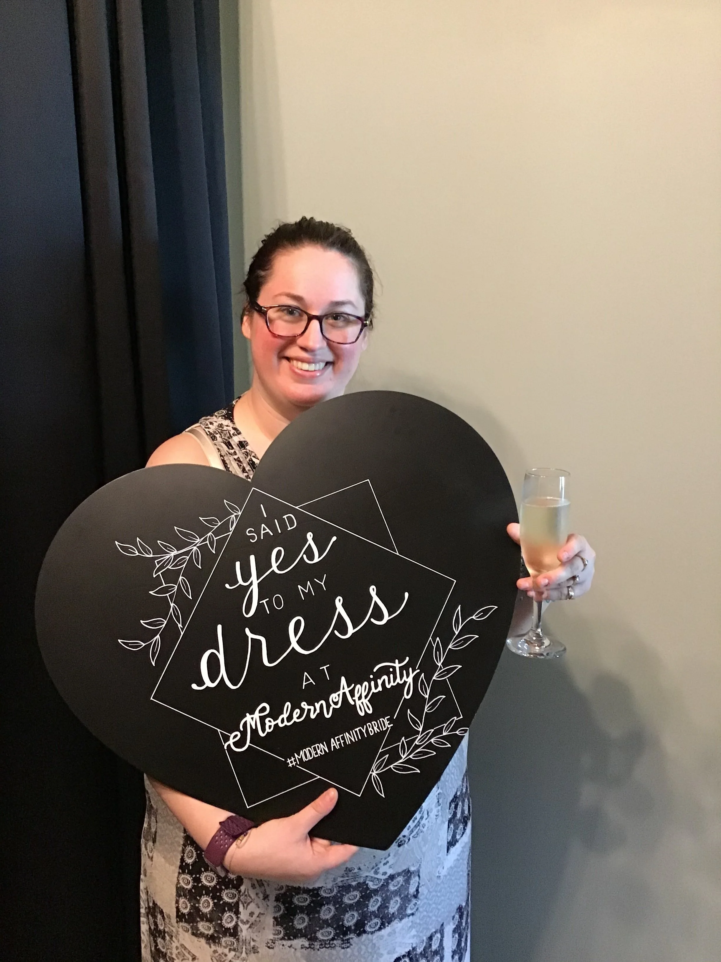 Woman holding a large heart-shaped sign that reads 'Said yes to my dress at Modern Affinity. #ModernAffinityBride,' while also holding a glass of champagne, smiling for the camera.