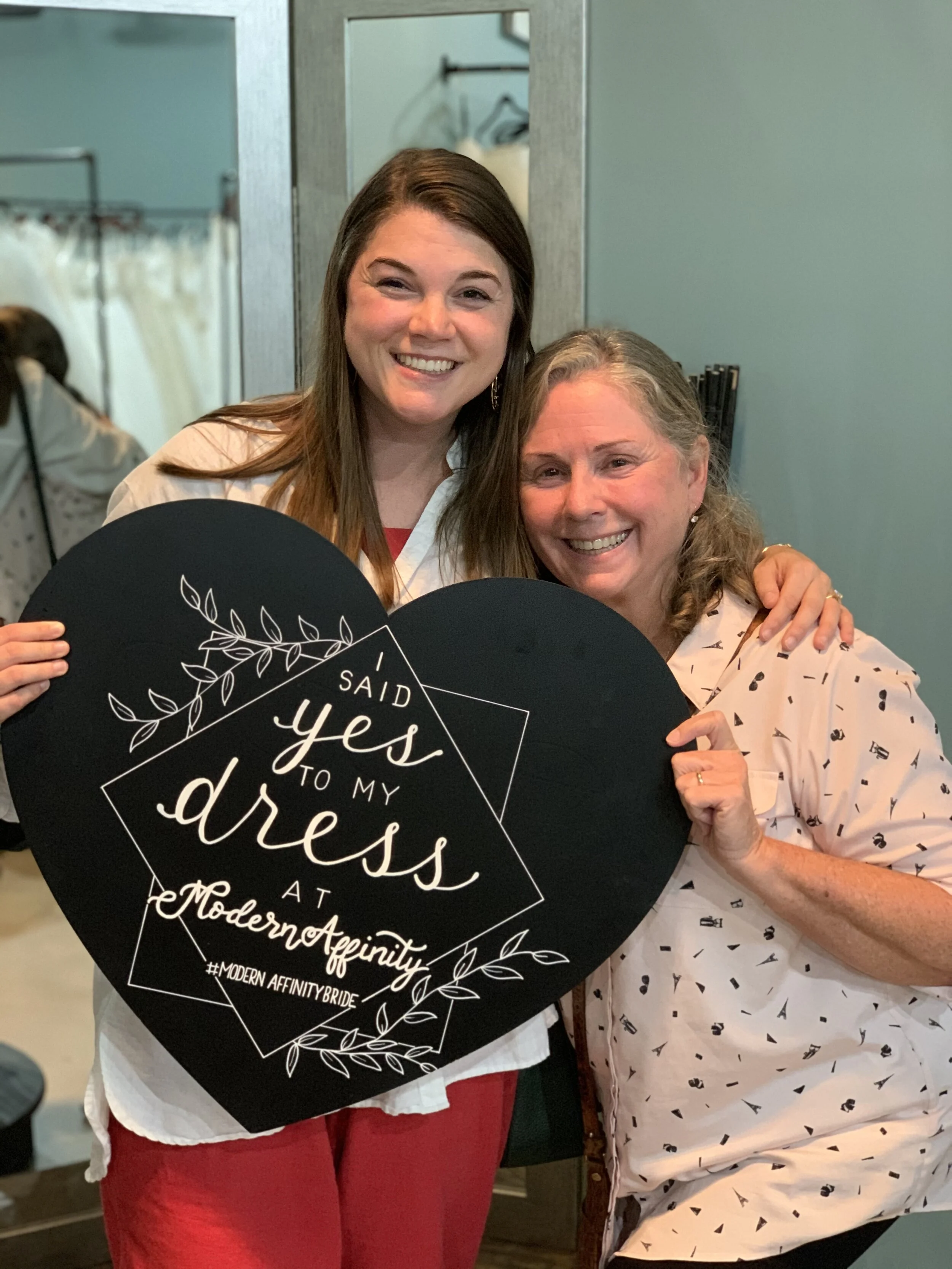 Two women smiling and holding a heart-shaped sign that reads, "I said yes to my dress at Modern Affinity."