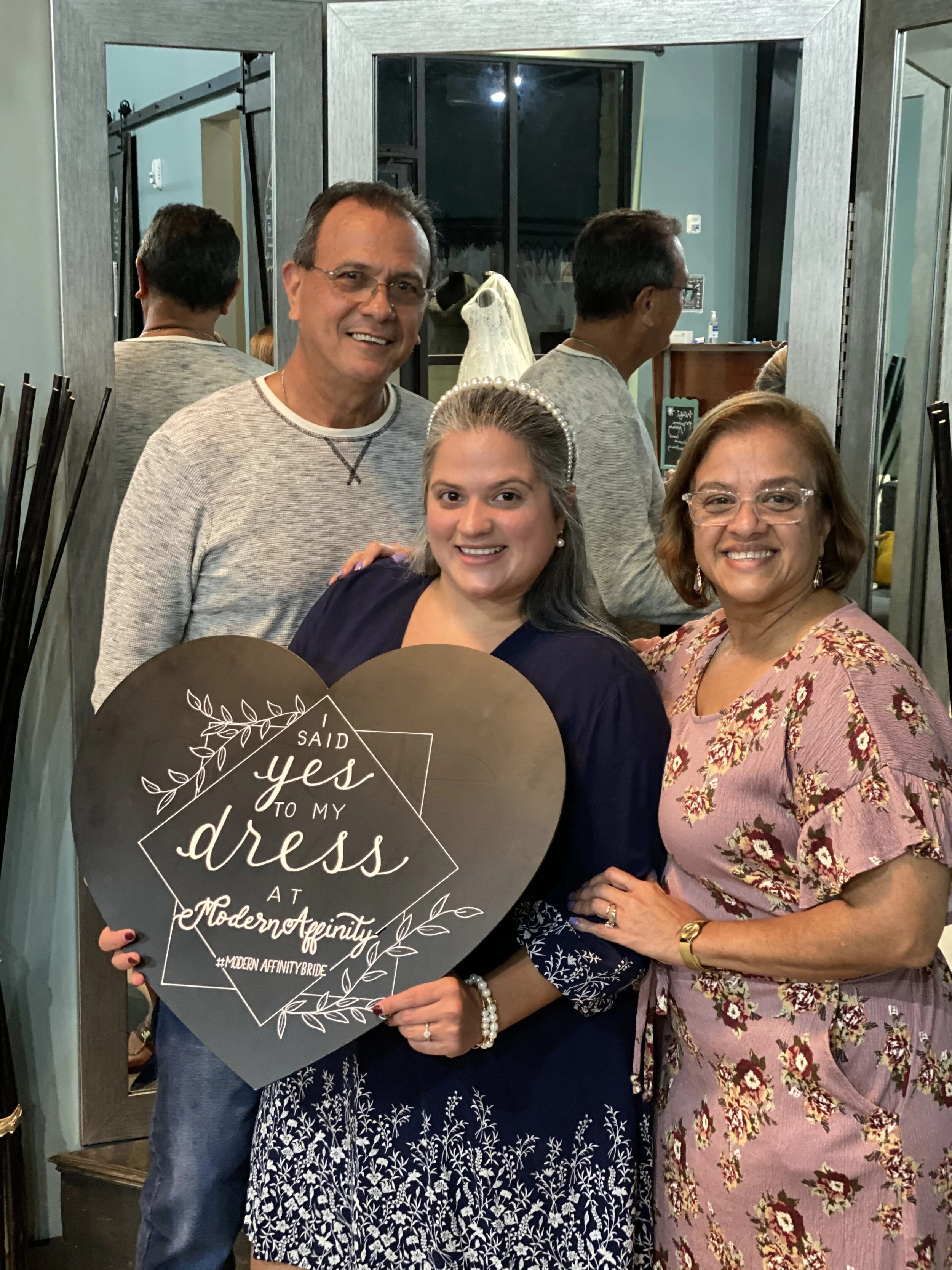 Three women and one man smiling at a bridal shower with a decorative heart-shaped sign that says 'I said yes to my dress at Modern Affinity'.