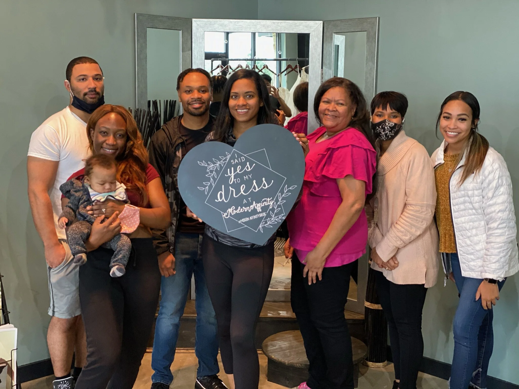 Group of eight people celebrating a wedding dress fitting at Modern Tapestry, with one holding a sign that says 'I said yes to my dress'
