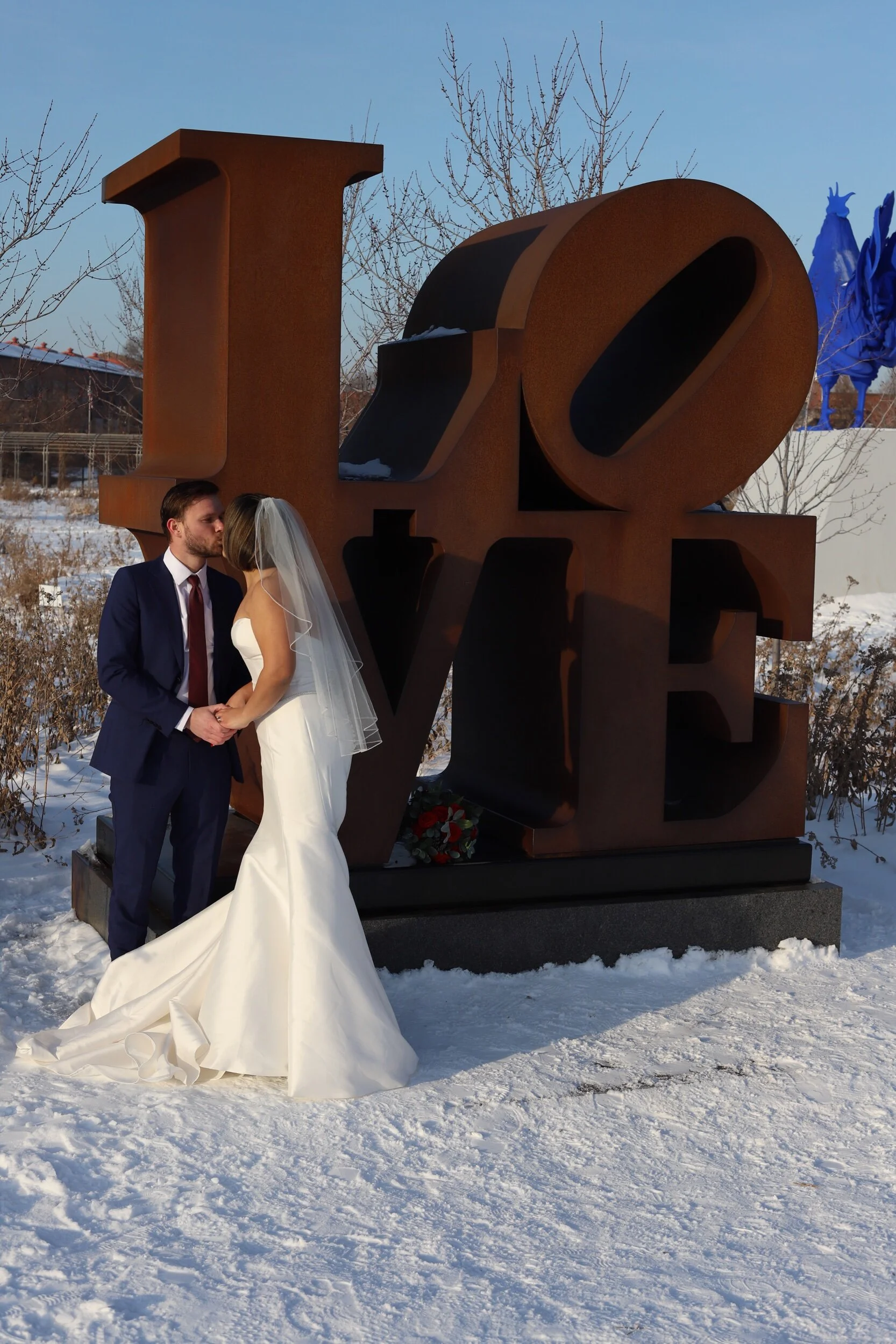 Bride and groom in wedding attire holding hands in front of a large LOVE sculpture in a snowy outdoor setting.