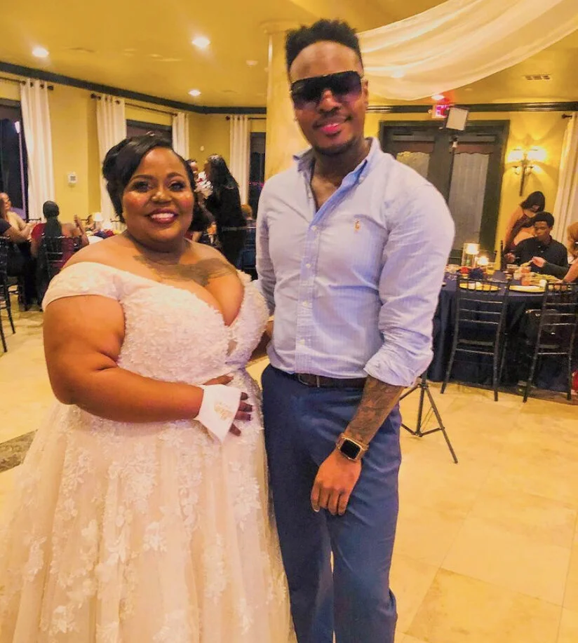 A woman in a light pink lace dress and a man in a light blue shirt and dark sunglasses standing together at a celebration event in a decorated indoor venue, with guests sitting at tables in the background.