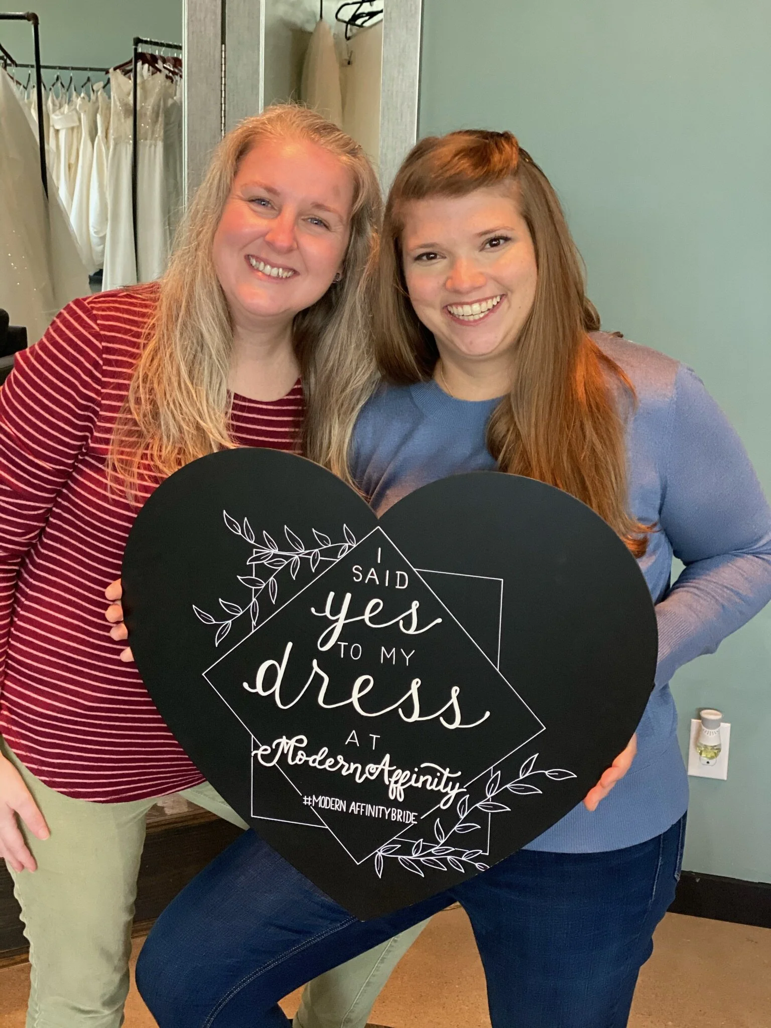 Two women smiling and holding a black heart-shaped sign that says, 'I said yes to my dress at Modern Affinity #ModernAffinityBride,' standing inside a room.