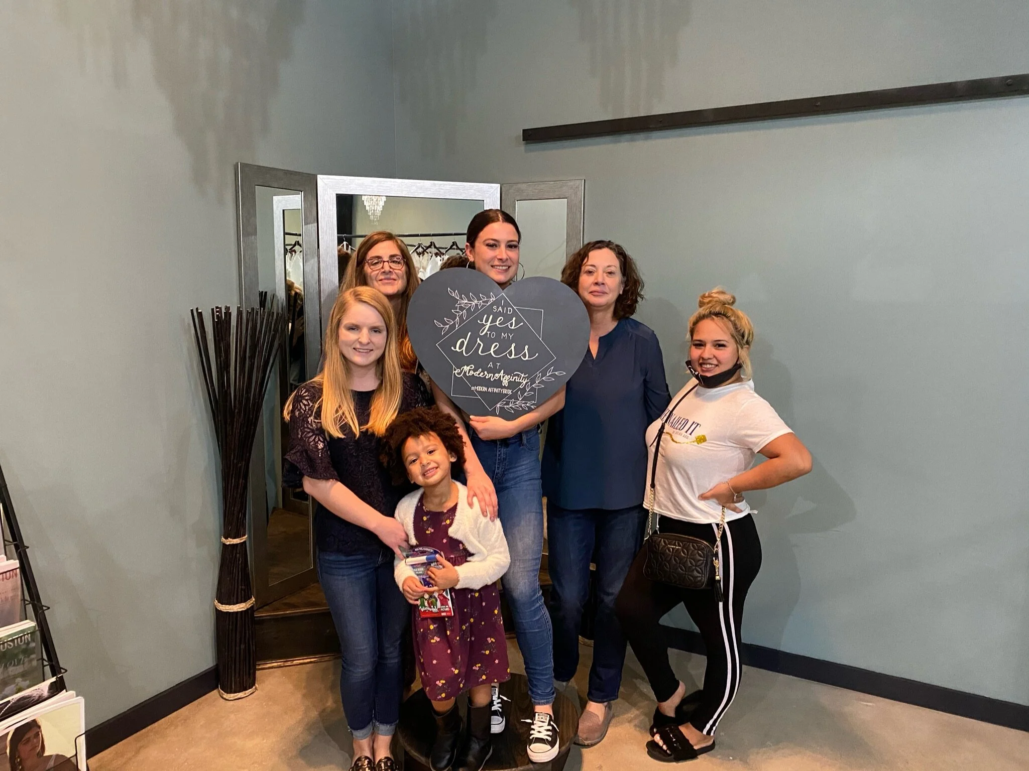 Seven women and one young girl posing together indoors for a photo. One woman is holding a heart-shaped sign that reads 'Said Yes to My Dress.' The group appears happy and is standing near a mirror and a coat rack.