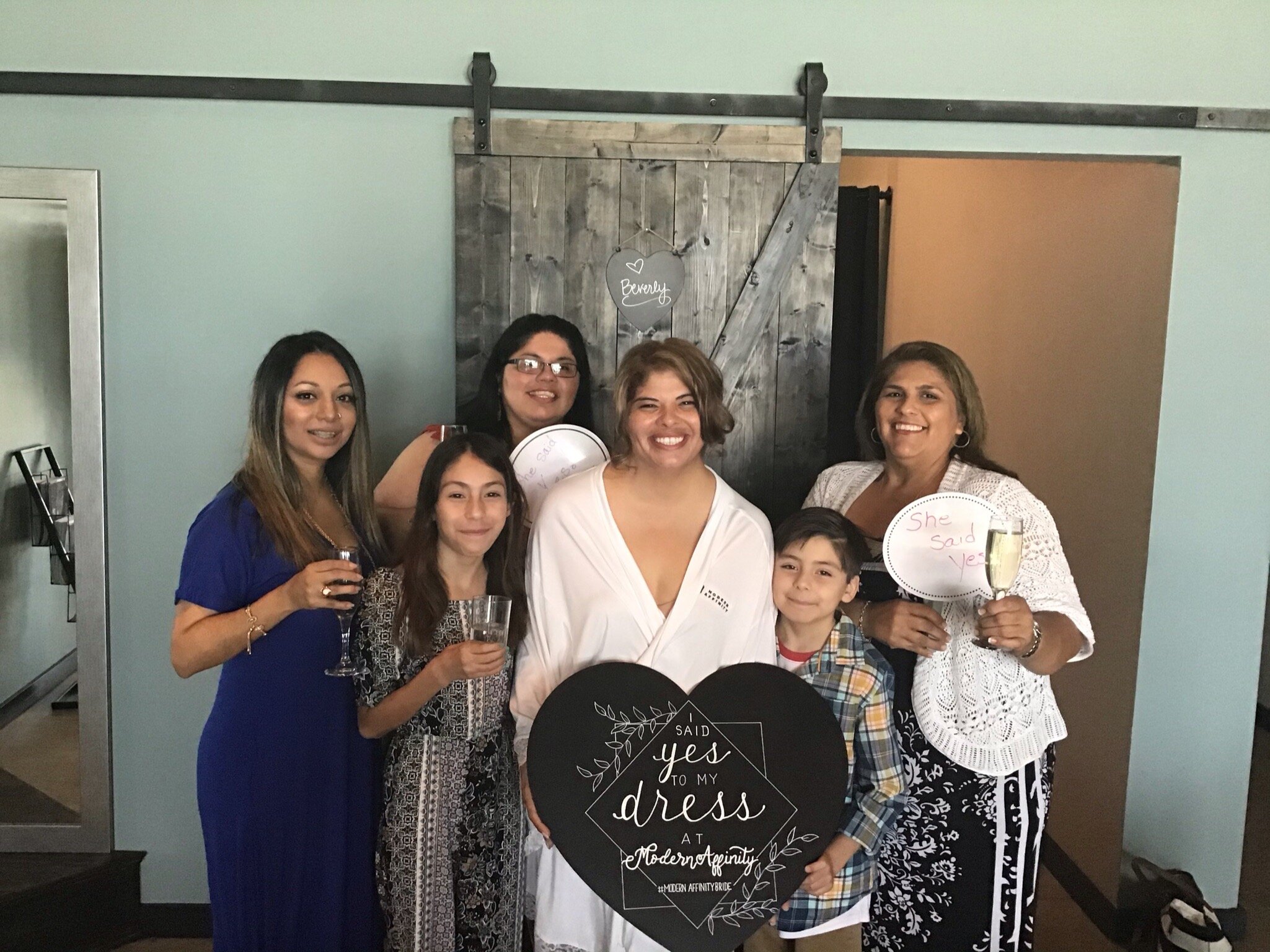 Group of women and children celebrating a wedding or engagement, holding drinks, with a bride in a white dress or robe, in front of a rustic wooden backdrop, some holding signs with celebratory messages.