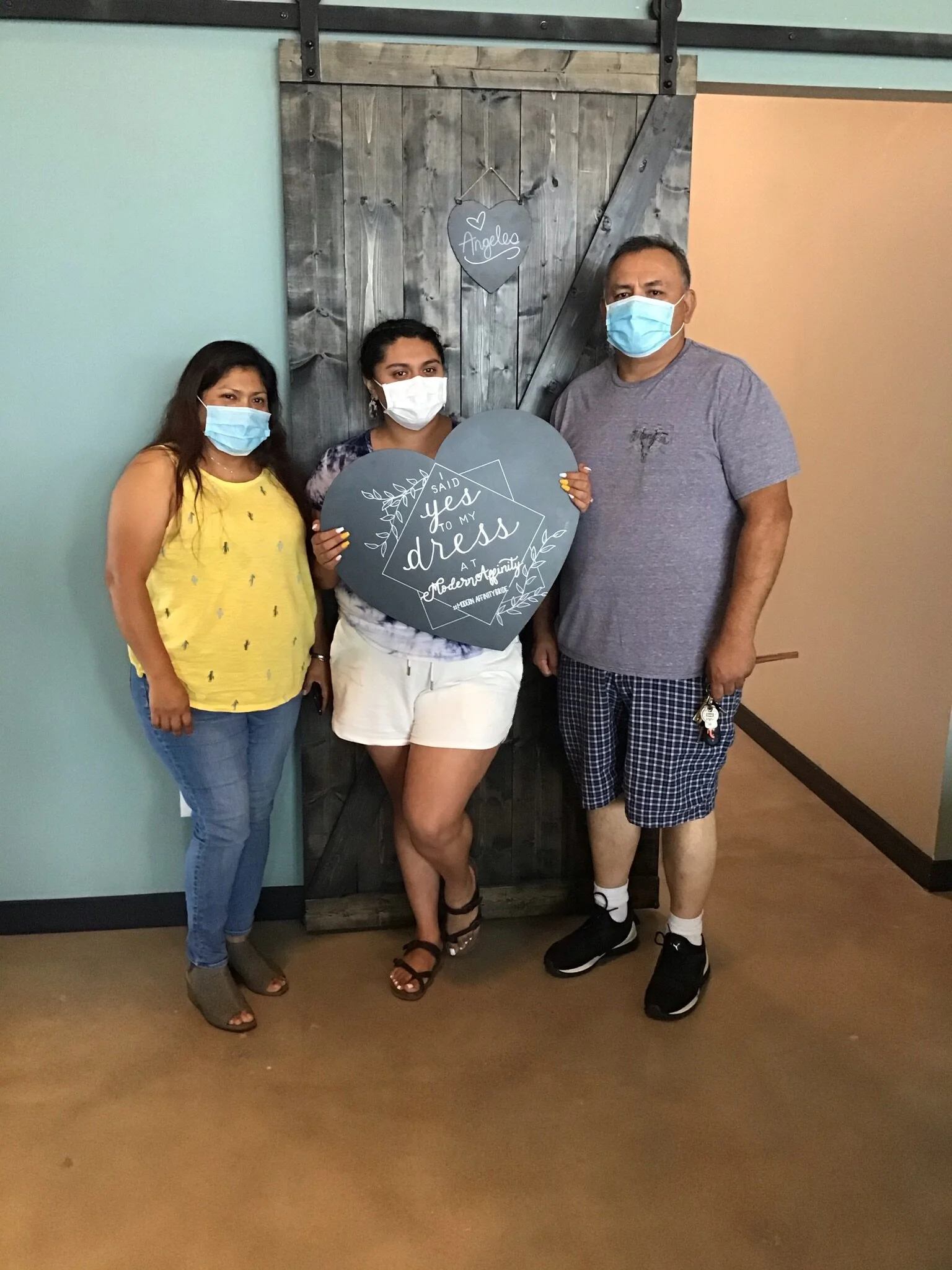 Three people wearing face masks standing indoors in front of a wooden sliding barn door, holding a large heart-shaped sign with the text 'Said yes to my dress at Modern Tuxedo.'