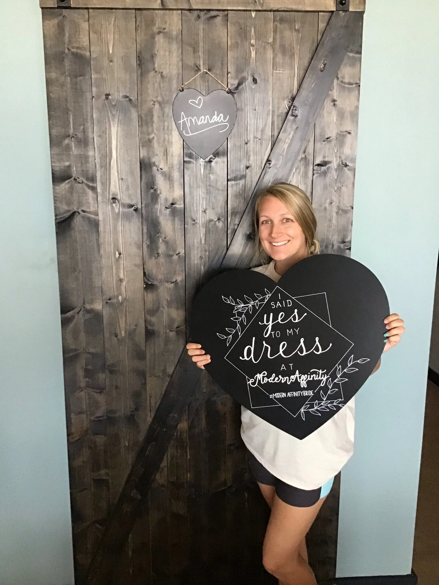 Woman holding a large heart-shaped sign that says "I said yes to my dress at Modern Affinity." She is standing in front of a rustic wooden door with a small chalkboard heart above that has "Amanda" written on it.