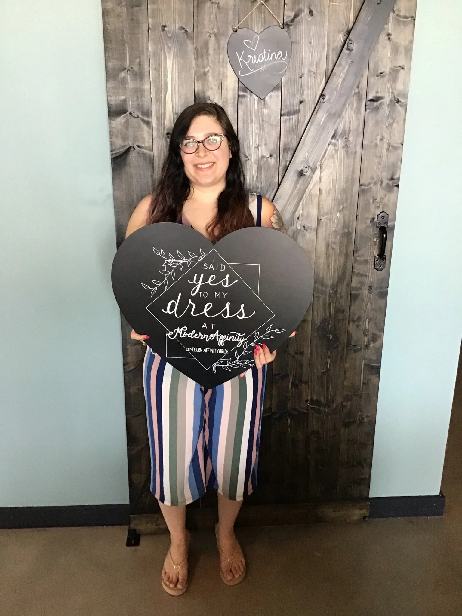 Woman with glasses holding a large black heart-shaped sign that reads 'I said yes to my dress at Modern Affinity' while standing in front of a rustic wooden door with a hanging heart-shaped sign that says 'Krustina'.