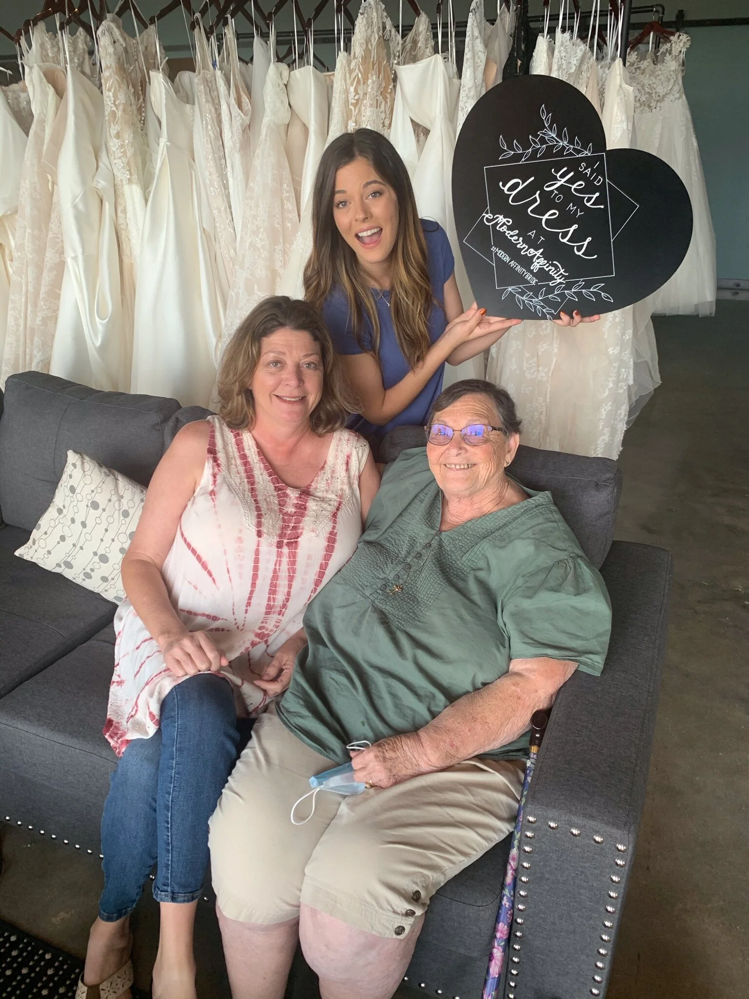 A young woman holding a black heart-shaped sign with white lettering that reads 'I said yes to my dress at Modern Bridal,' standing behind an older woman and man sitting on a gray sofa, with wedding dresses hanging in the background.