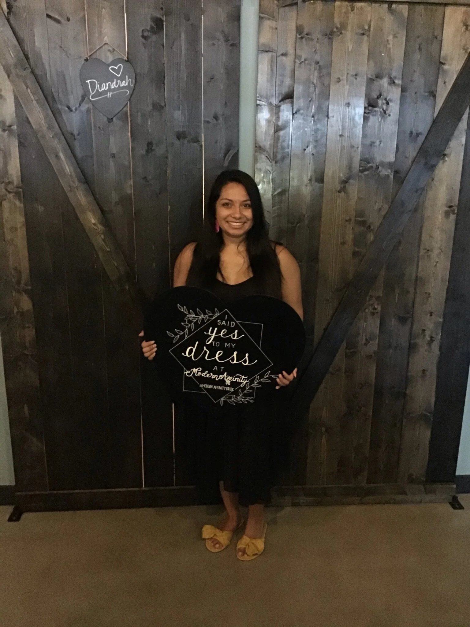 A woman in black dress and yellow shoes standing in front of a wooden barn door, holding a black sign that reads 'I said yes to my dress at Modern Affinity.' There is a heart-shaped chalkboard sign on the wall with the name 'Diandrah' written on it.