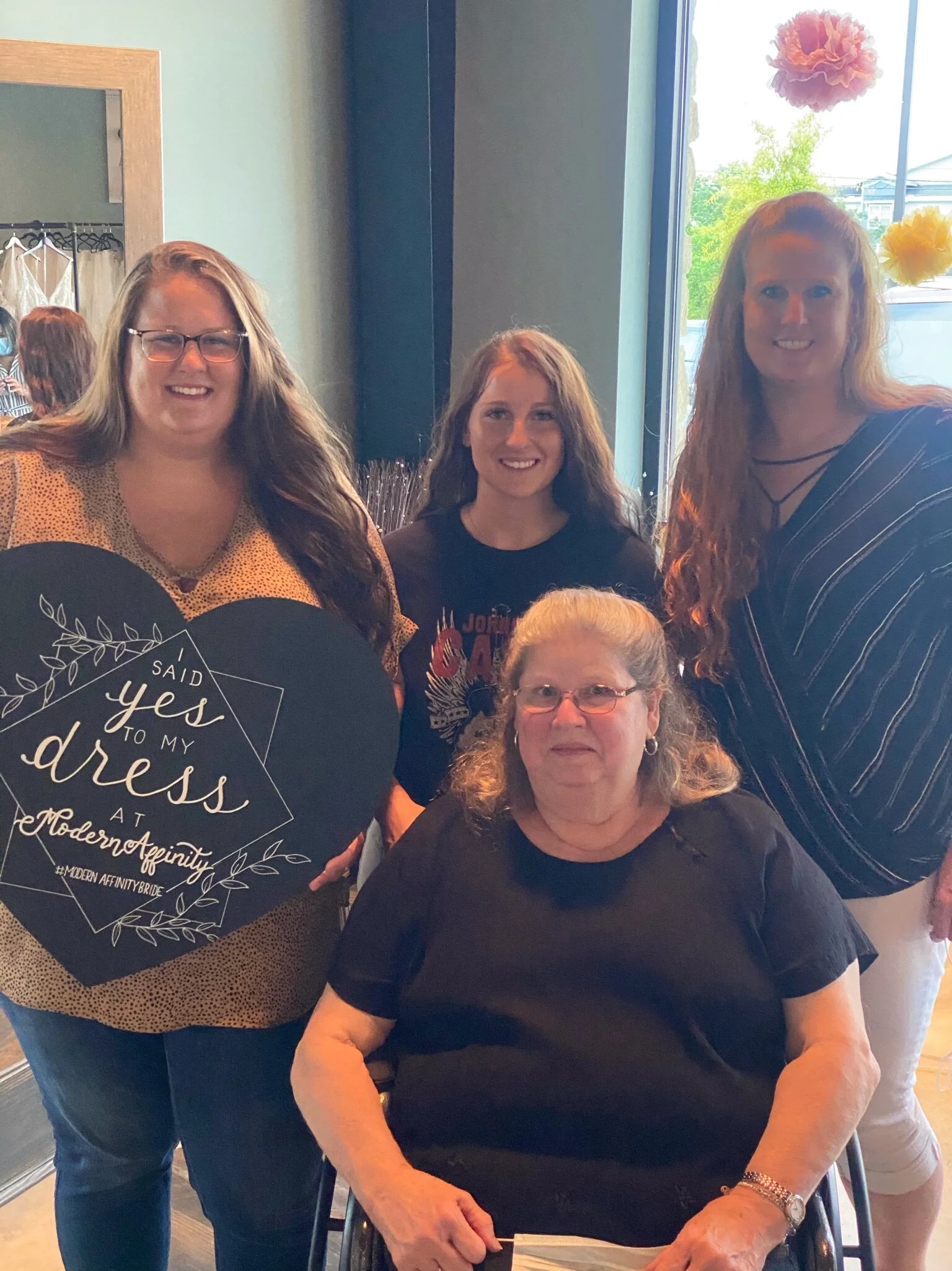 Four women gathered together, with one seated in a wheelchair, inside a room with large windows and colorful decorations hanging from the ceiling.
