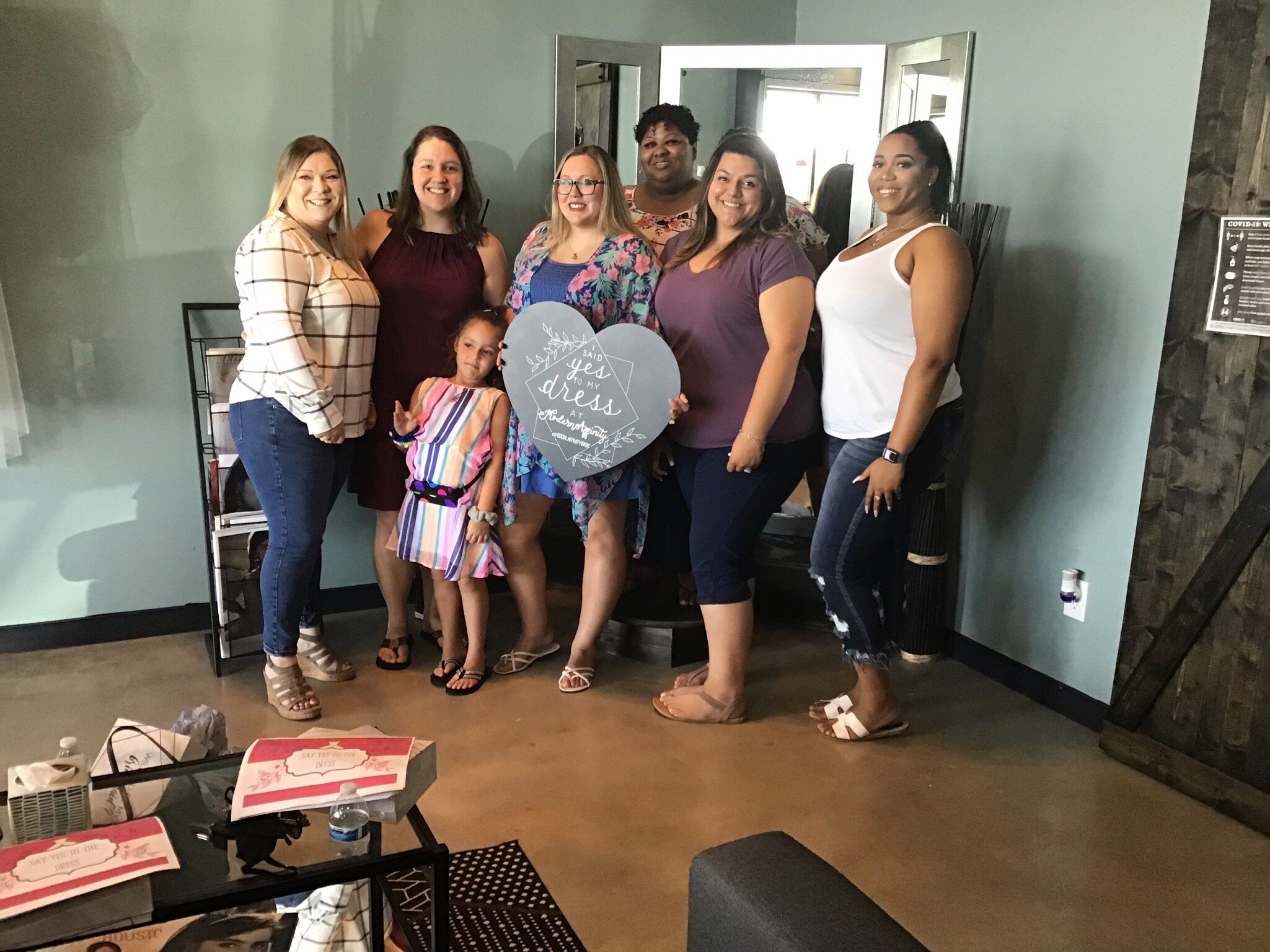 A group of seven women and one young girl posing indoors, smiling, with the women holding a heart-shaped sign that reads 'Said yes to my dress and adventures'.