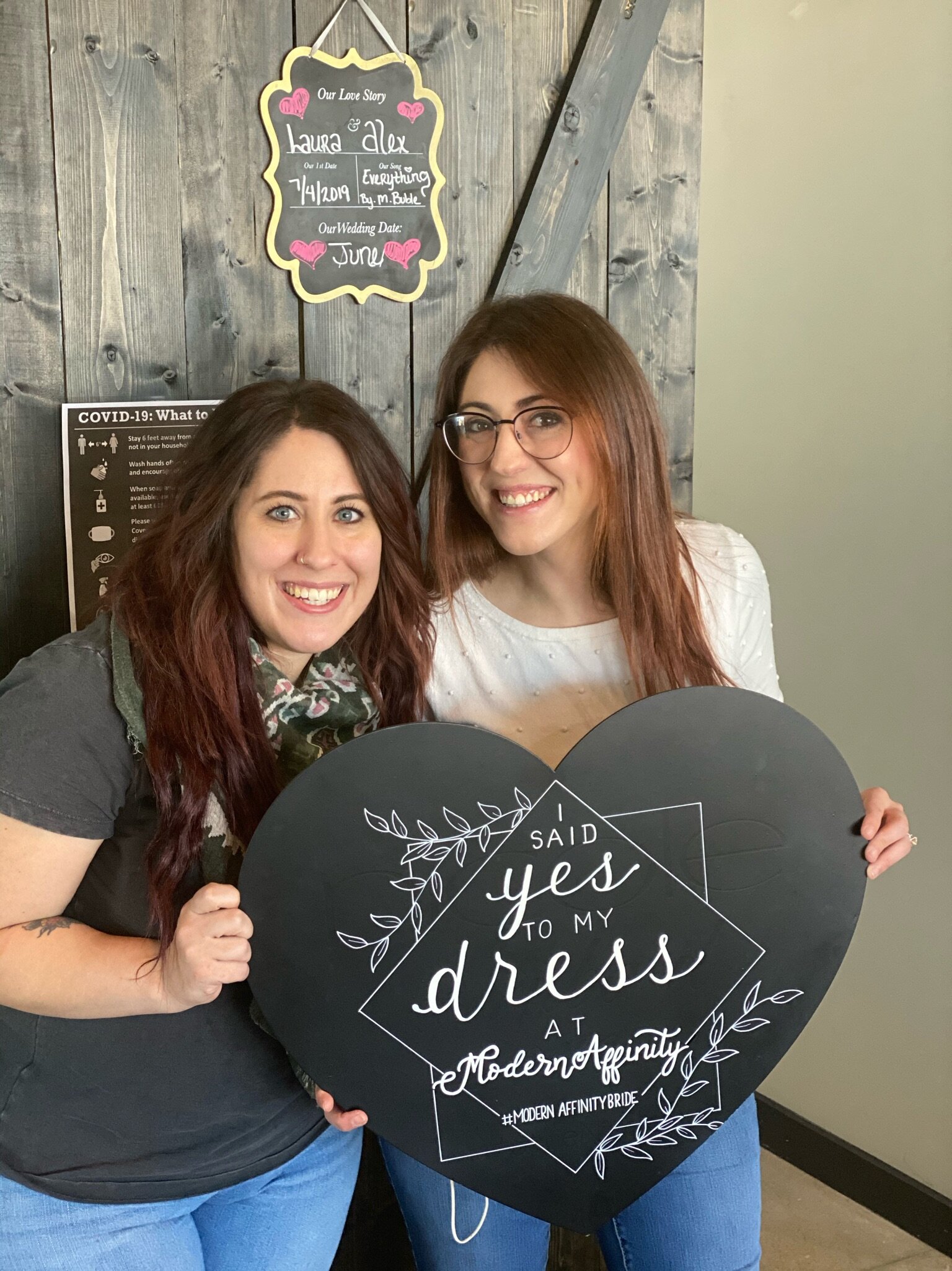 Two women smiling, holding a large black heart-shaped sign with white text that reads "I said yes to my dress at Modern Affinity." They are standing indoors, with a wooden wall and a decorative board behind them.