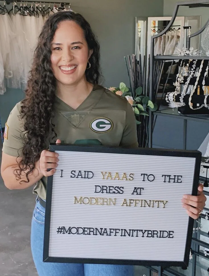 A young woman with long, curly hair and a big smile, wearing a Green Bay Packers jersey, standing inside a room and holding a sign with a humorous message about her wedding dress, mentioning 'Modern Affinity' and a hashtag #MODERNAFFINITYBRIDE.