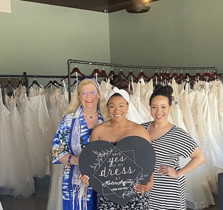 Three women standing together in a bridal boutique with wedding dresses hanging behind them. The woman in the middle is holding a black heart-shaped sign that says, 'Said yes to my dress at Modern Promises.'