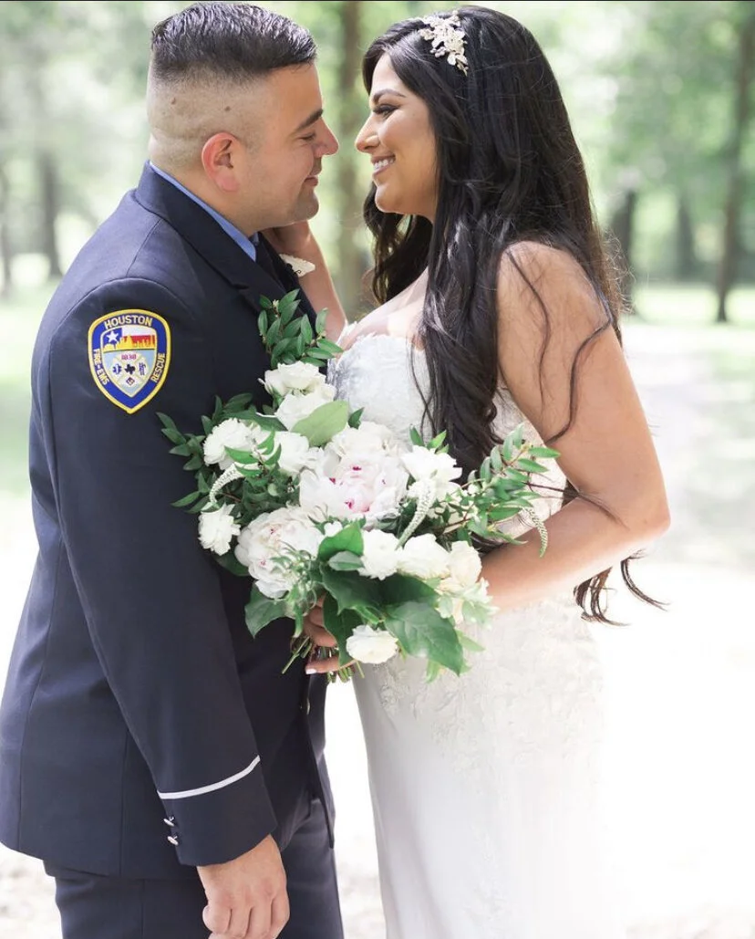 A bride and groom in a close, happy moment outdoors, with the groom in a police uniform and the bride holding a large bouquet of white flowers.