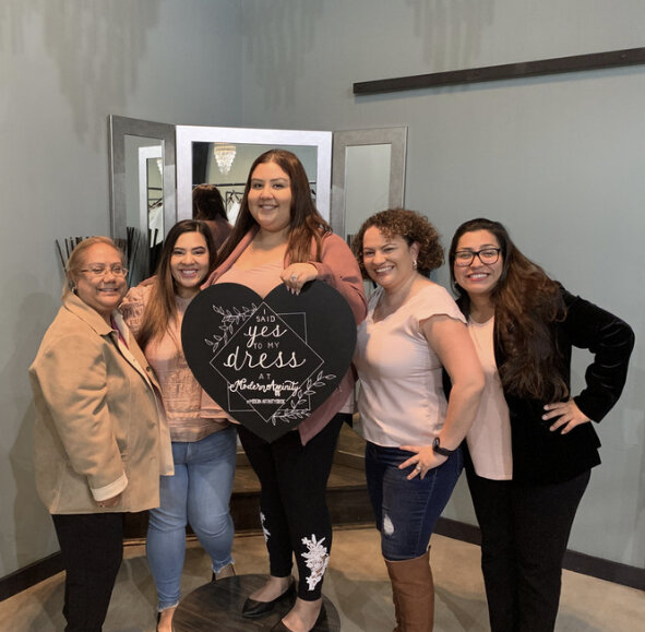 Five women standing together inside a room, smiling, with one holding a sign that says 'said YES to my dress at grandpappy's'.