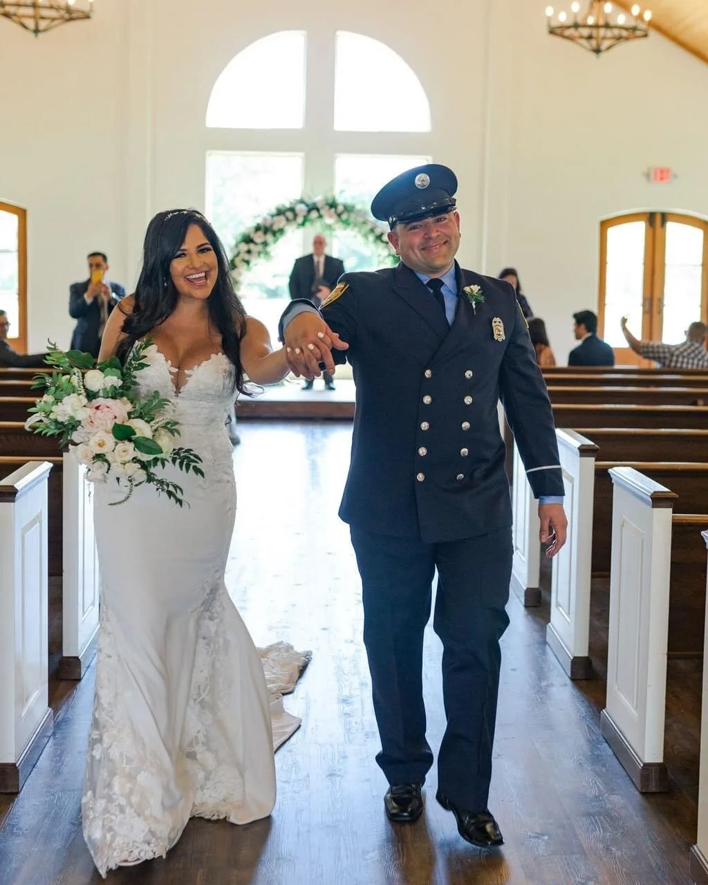 A bride in a white wedding dress holding a bouquet of white flowers and greenery, smiling, walking down the aisle with a police officer in uniform inside a church with guests in the background, some taking photos.