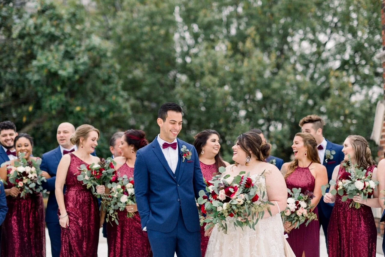 A wedding group photo featuring a bride and groom with their wedding party outdoors, surrounded by trees. The bride wears a white gown and holds a bouquet, while the groom wears a blue suit with a burgundy bowtie. The bridesmaids wear matching burgun
