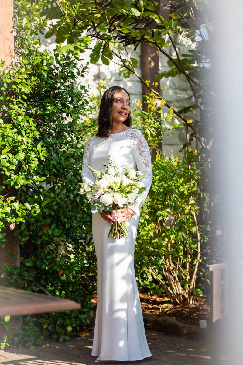A woman in a white wedding dress holding a bouquet of white flowers, standing outdoors surrounded by green foliage and sunlight.