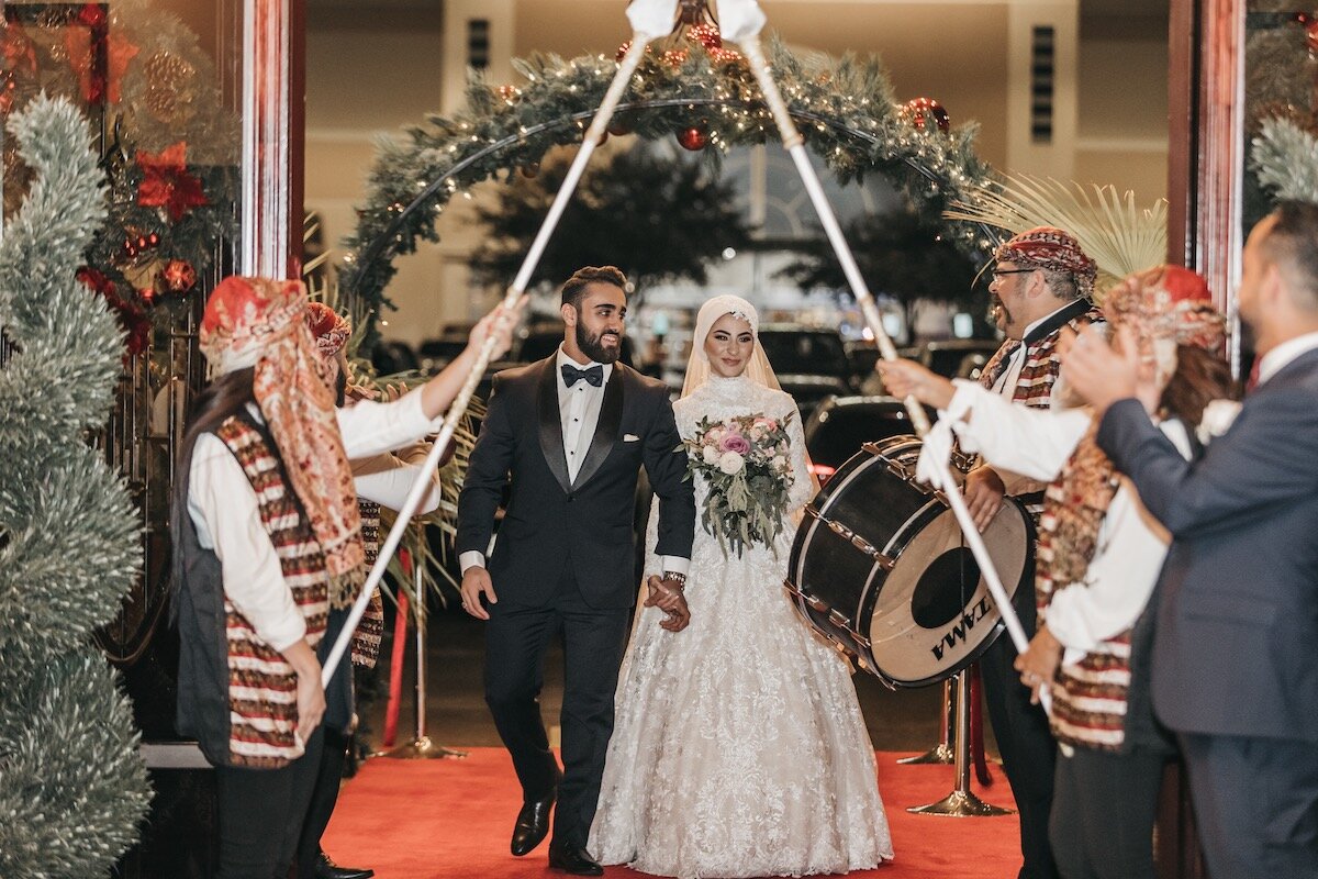 Bride and groom walking through a decorated archway surrounded by musicians in traditional attire at a wedding reception.