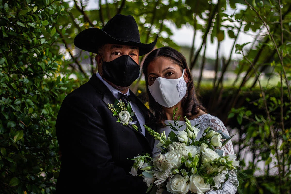 A couple dressed in wedding attire, both wearing black and white face masks, standing close together outdoors among green foliage, with the man wearing a black cowboy hat and a boutonnière, and the woman holding a bouquet of white flowers.