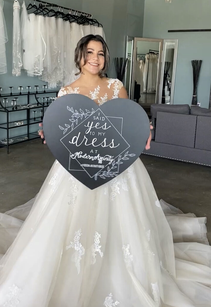 A bride in a wedding gown holding a heart-shaped sign that reads, 'I said yes to my dress at Modern Makeup,' standing in a bridal boutique with dresses, mirrors, and furniture in the background.