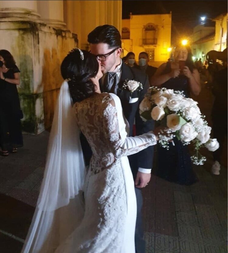 Wedding couple kissing outside at night, the bride wearing a lace wedding dress with long sleeves and veil, holding a bouquet of white flowers, and the groom in a dark suit with glasses, surrounded by guests taking photos.