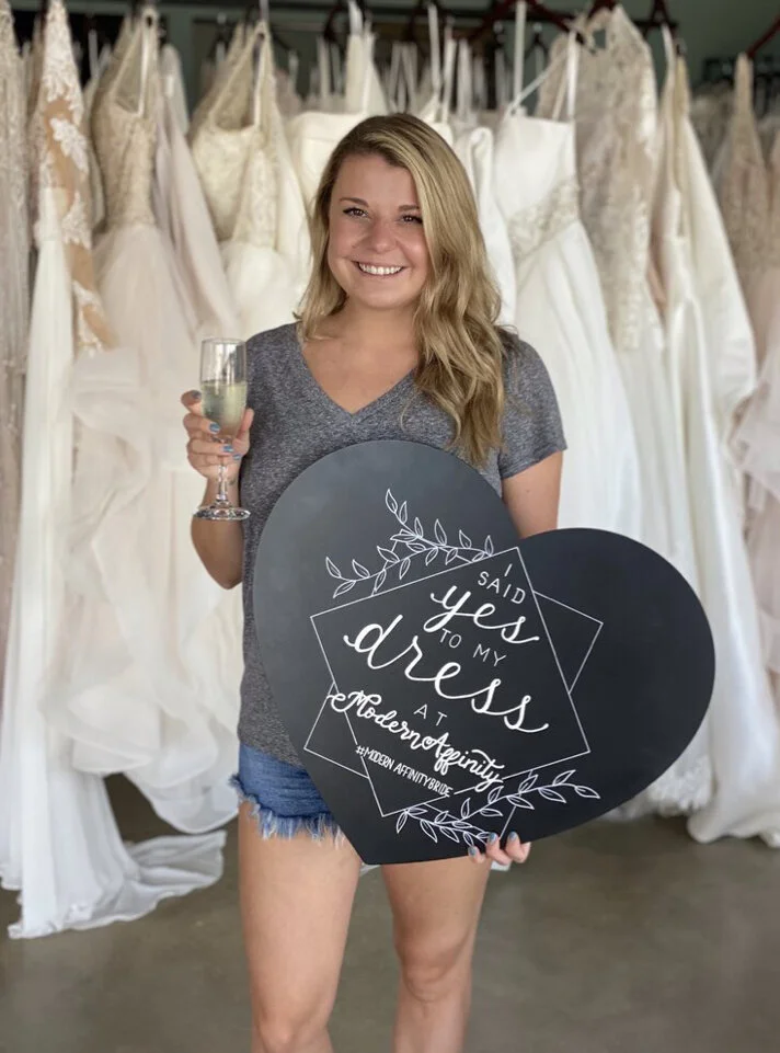 A smiling woman holding a glass of champagne and a heart-shaped sign with the text "Said Yes To My Dress At Modern Affinity" stands in front of wedding dresses.