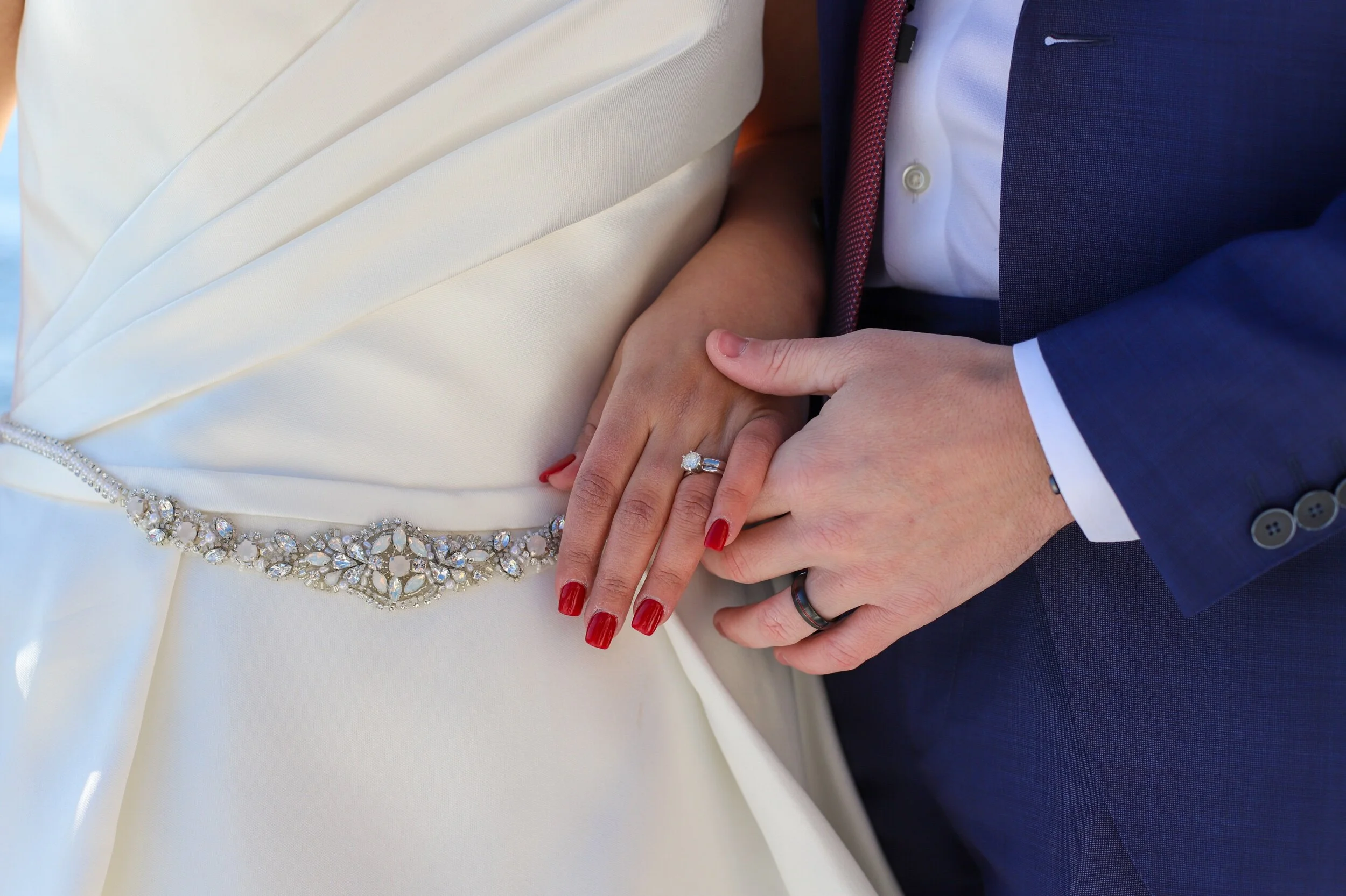 Close-up of a bride and groom holding hands, showing wedding rings, with the bride wearing a white dress with beaded embellishments and red nail polish, and the groom in a navy suit.
