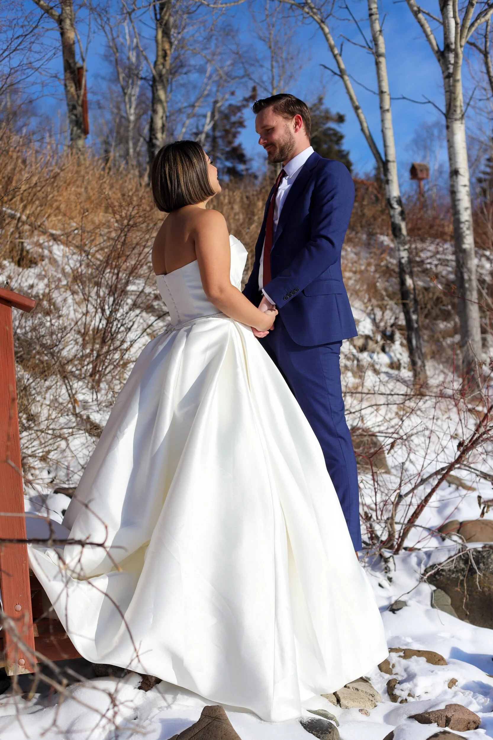 A bride and groom holding hands and standing together outdoors on a snowy hillside, with leafless trees and blue sky in the background.