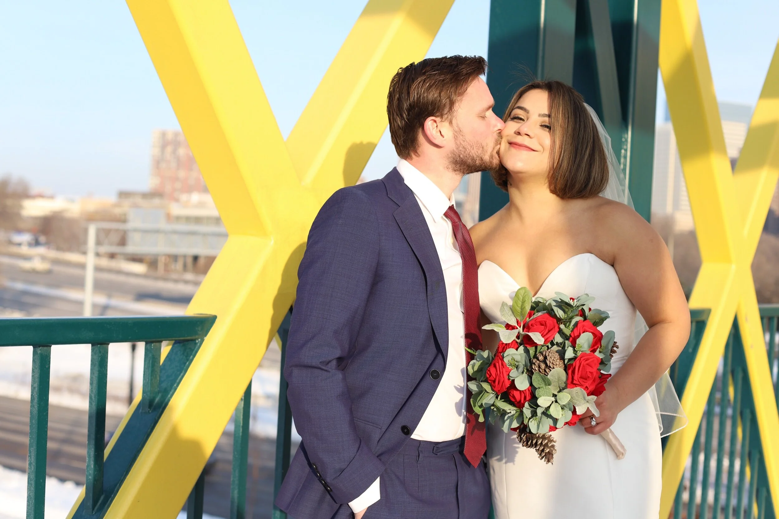 A bride and groom stand on a bridge, with the groom kissing the bride's cheek. The bride is smiling and holding a bouquet of red roses, eucalyptus, and pinecones. The bride is wearing a strapless white wedding dress with a veil, and the groom is wear