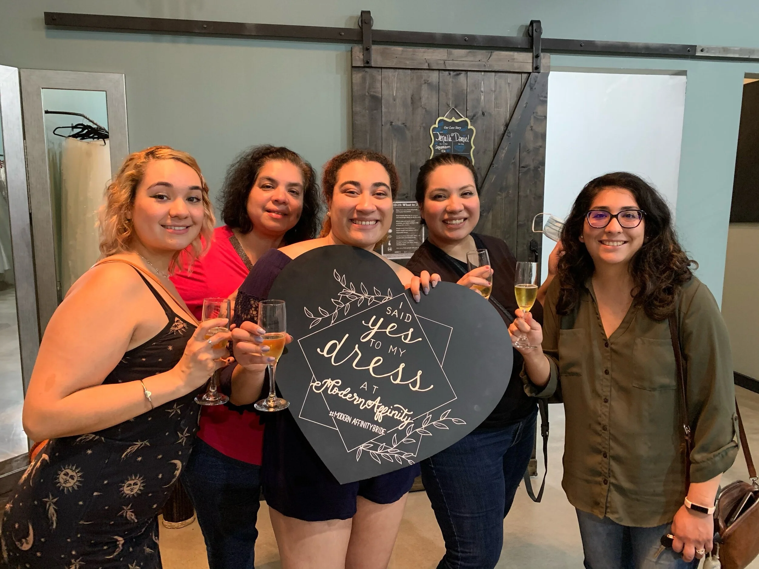 Five women celebrating at a bridal shower, holding glasses of champagne around a large black circular sign that reads "Said Yes to My Dress at Modern Affinity". They are smiling and dressed casually.