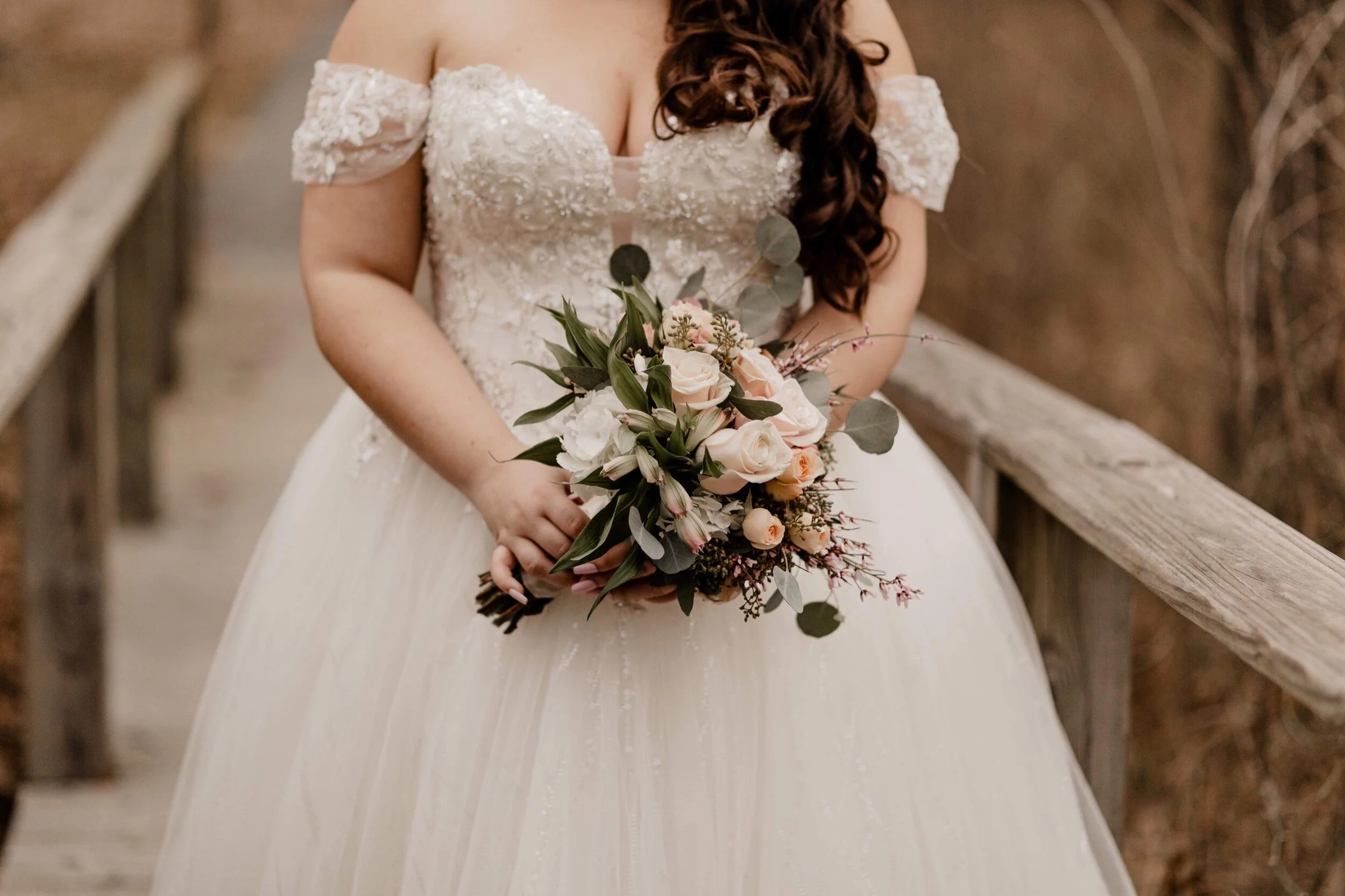 A woman in a wedding dress holding a bouquet of pink and white flowers with green leaves, standing on a wooden bridge.