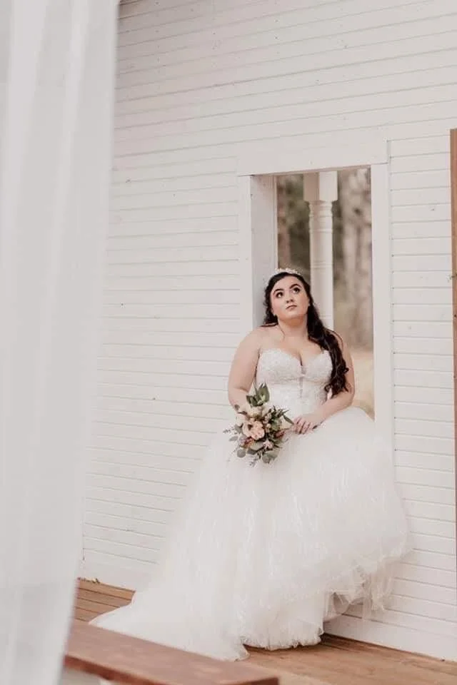 A bride in a white wedding dress holding a bouquet of flowers standing near a window in a white wooden house, looking contemplative.