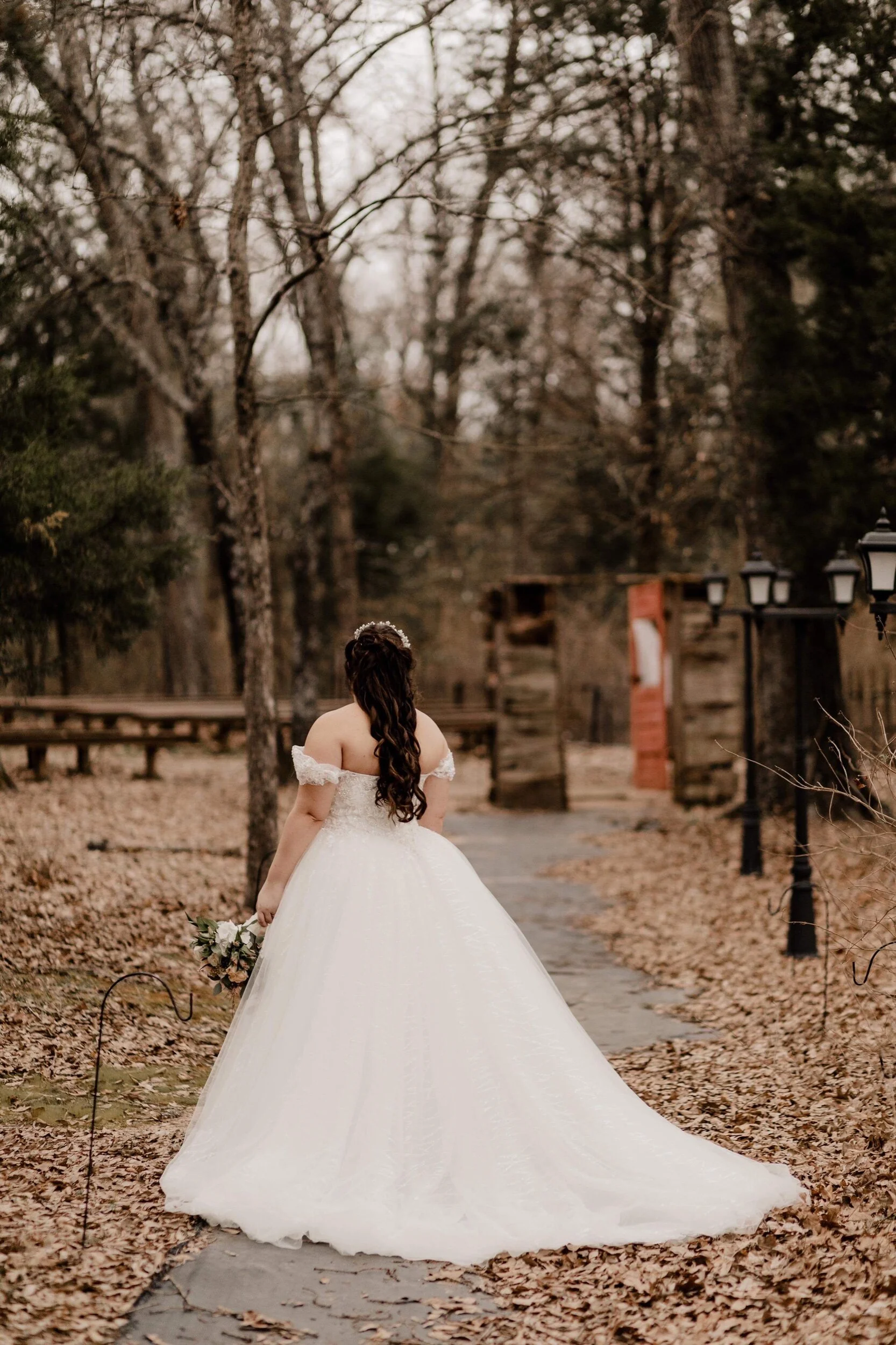 A bride in a white wedding gown holding a bouquet stands with her back to the camera on a tree-lined pathway covered with fallen leaves.