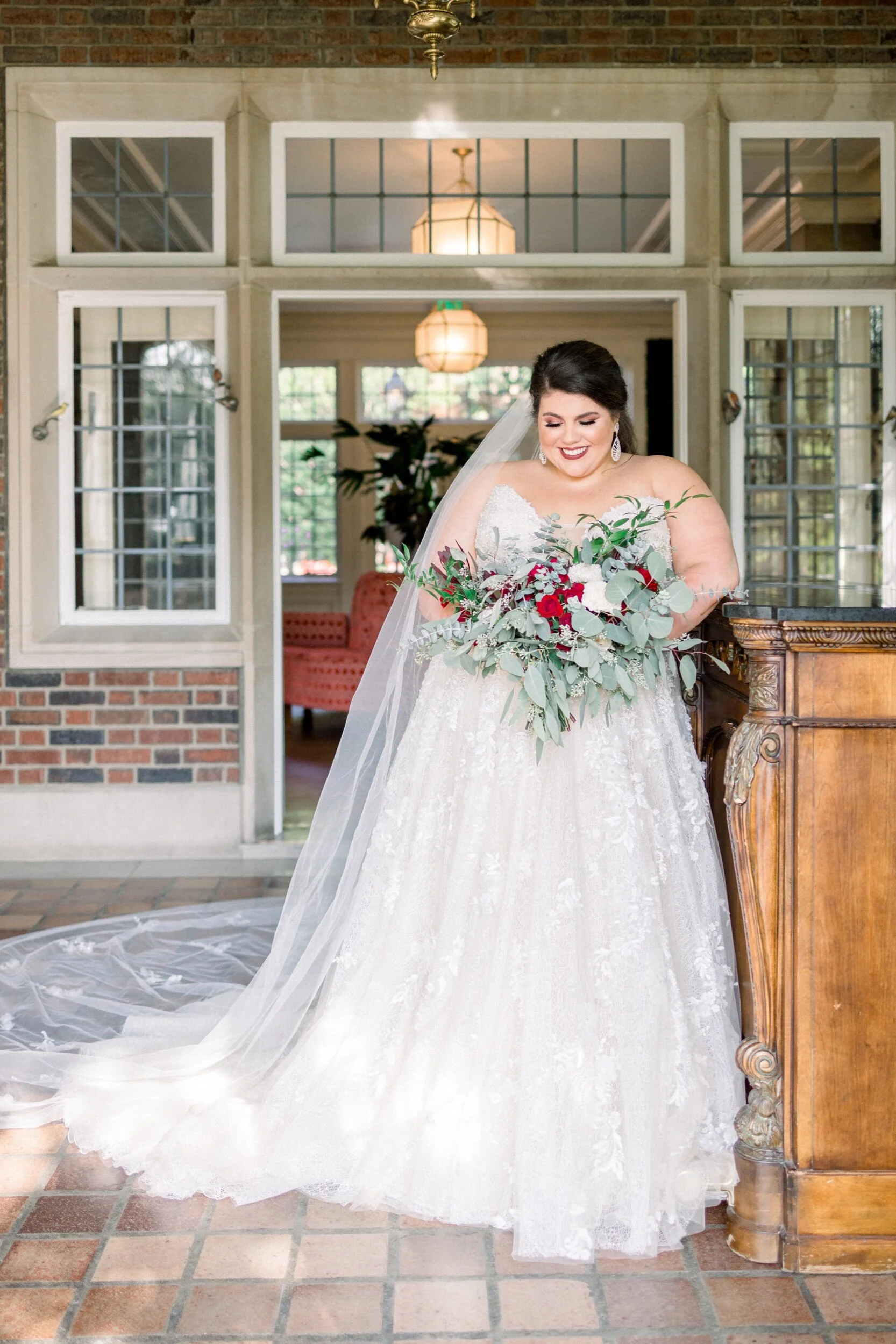 Bride in a white lace wedding gown holding a large bouquet of red and white flowers with green foliage, standing indoors with a brick and glass-paneled background.