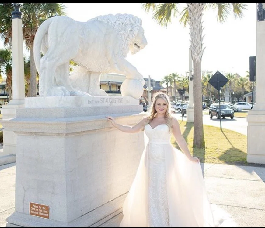 A woman in a wedding dress standing next to a large white lion statue in a plaza with palm trees and cars in the background.