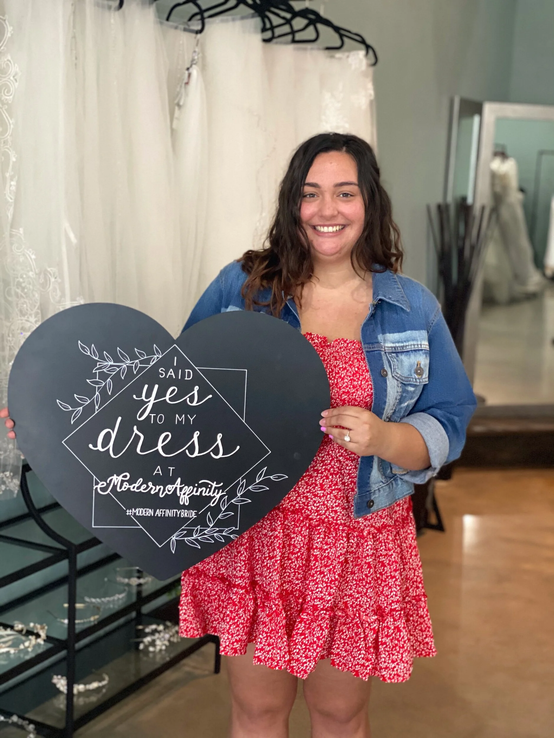Woman smiling while holding a large heart-shaped sign that reads 'I said yes to my dress at Modern Affinity' with decorative leaves and the hashtag '#ModernAffinityBride'. She is wearing a red dress with small white flowers and a denim jacket, standi