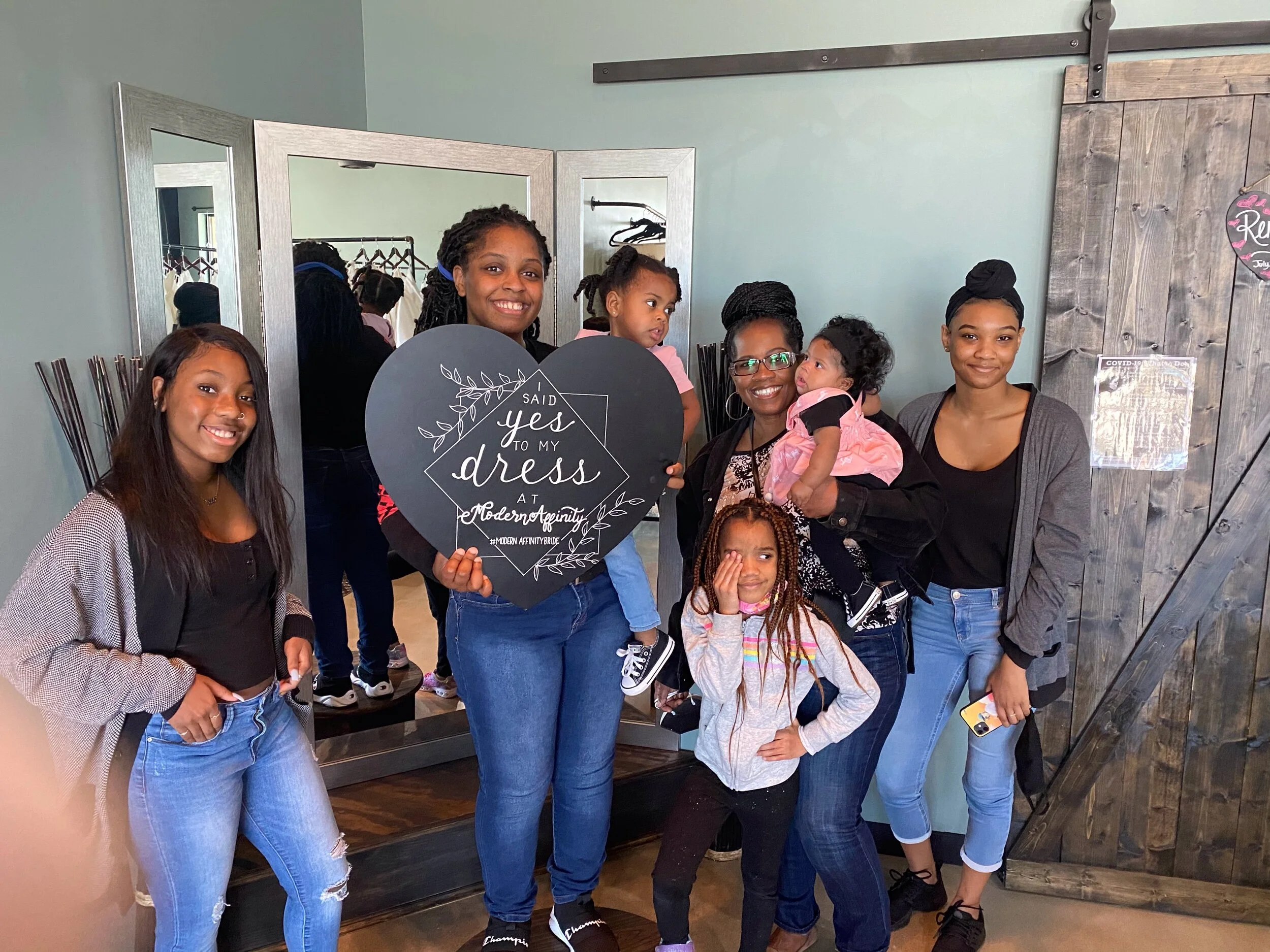 Group of seven women and girls posing in front of a mirror, with one woman holding a sign that says, 'Said yes to my dress at Modern Affinity.'